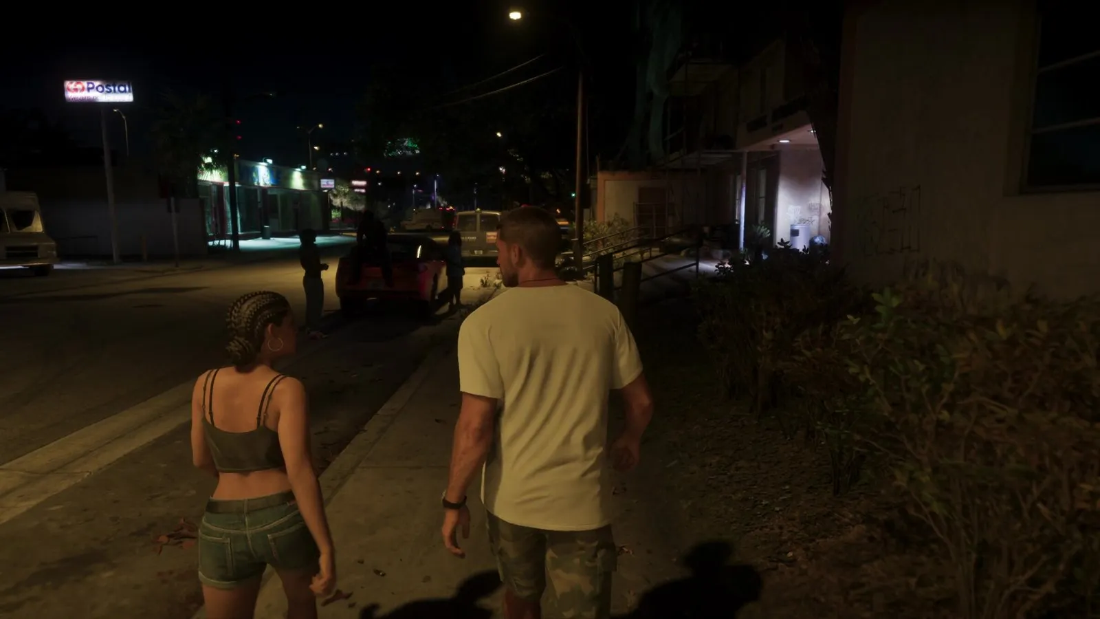 Two people walk along a night sidewalk while an ambulance with red roof lights sits further down the street past a parked van.