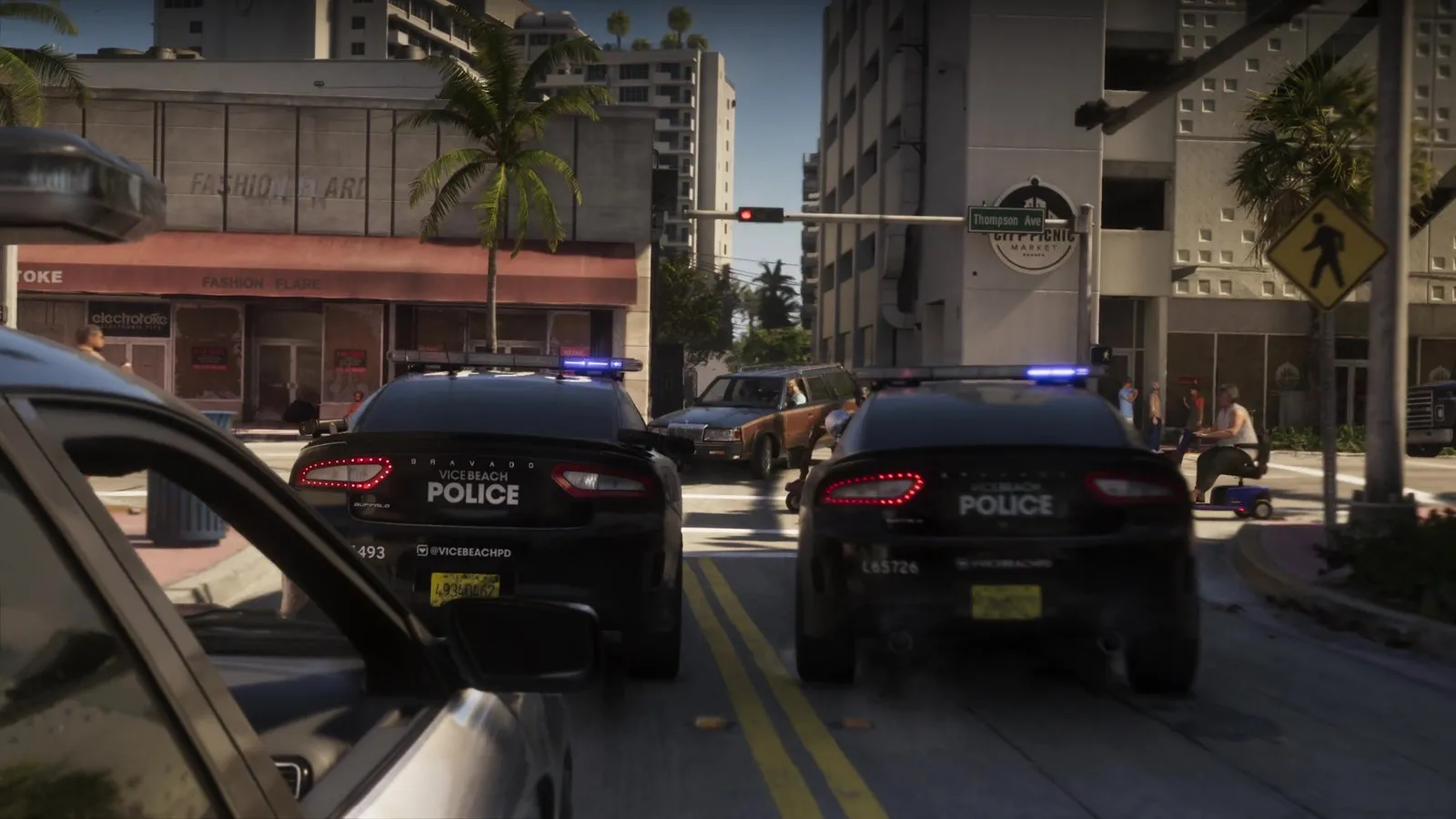 Two police cars with blue lights on reach an intersection as a wood paneled station wagon crosses in front of them.