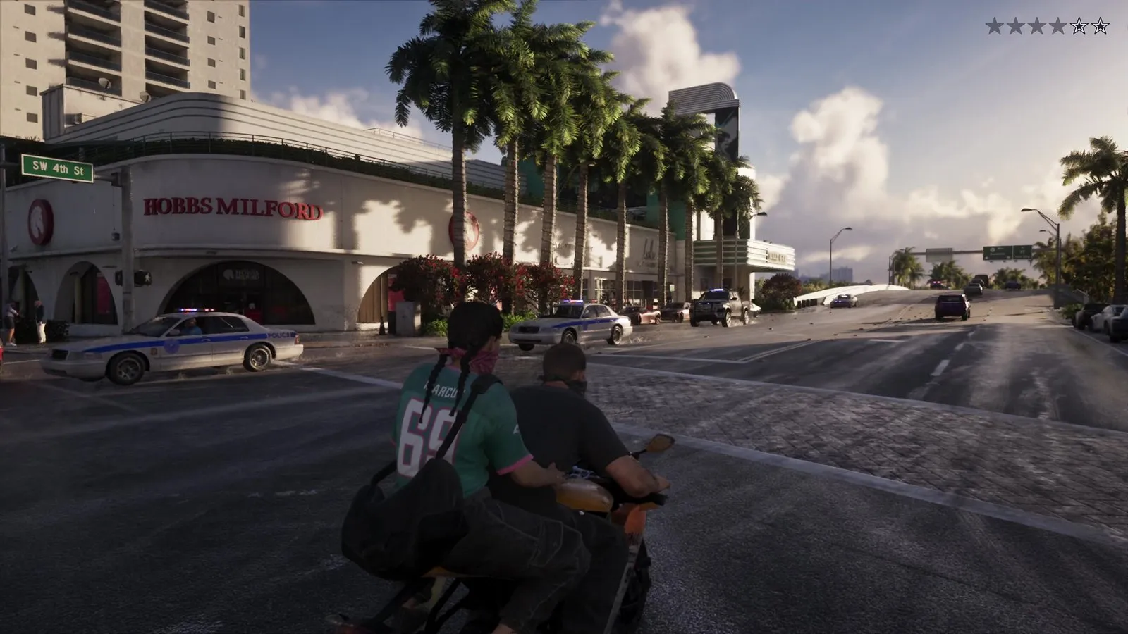 Two masked riders on a motorcycle stop at an intersection as police sedans and a pickup with lights on close in.