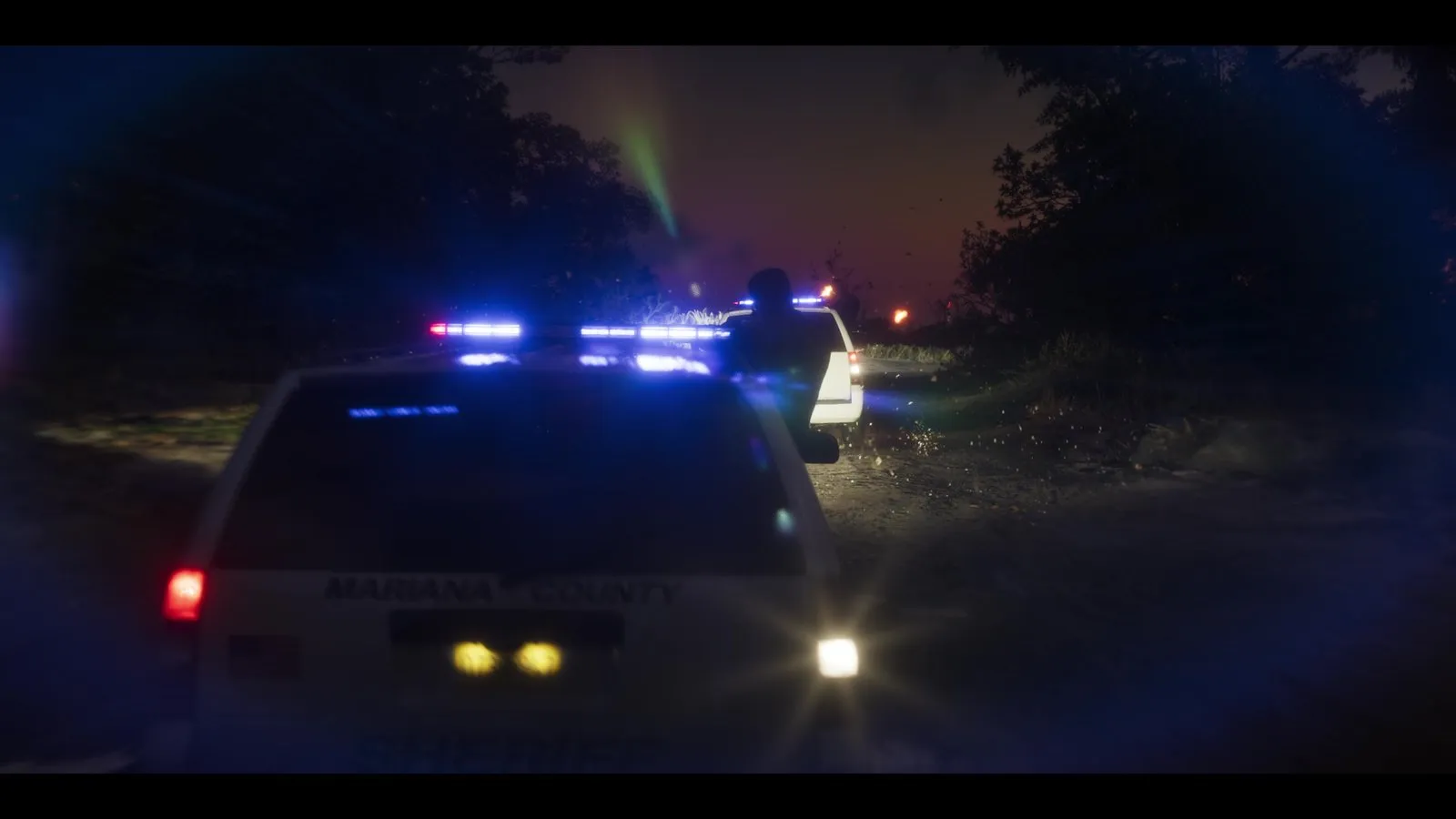 A sheriff SUV with lights flashing sits behind another police SUV on a dark wooded road, with small fires and light beams ahead.