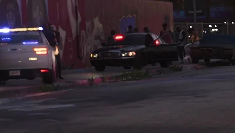 At dusk, a black sedan is parked at the curb behind a white police car, its sides dark with no markings visible.