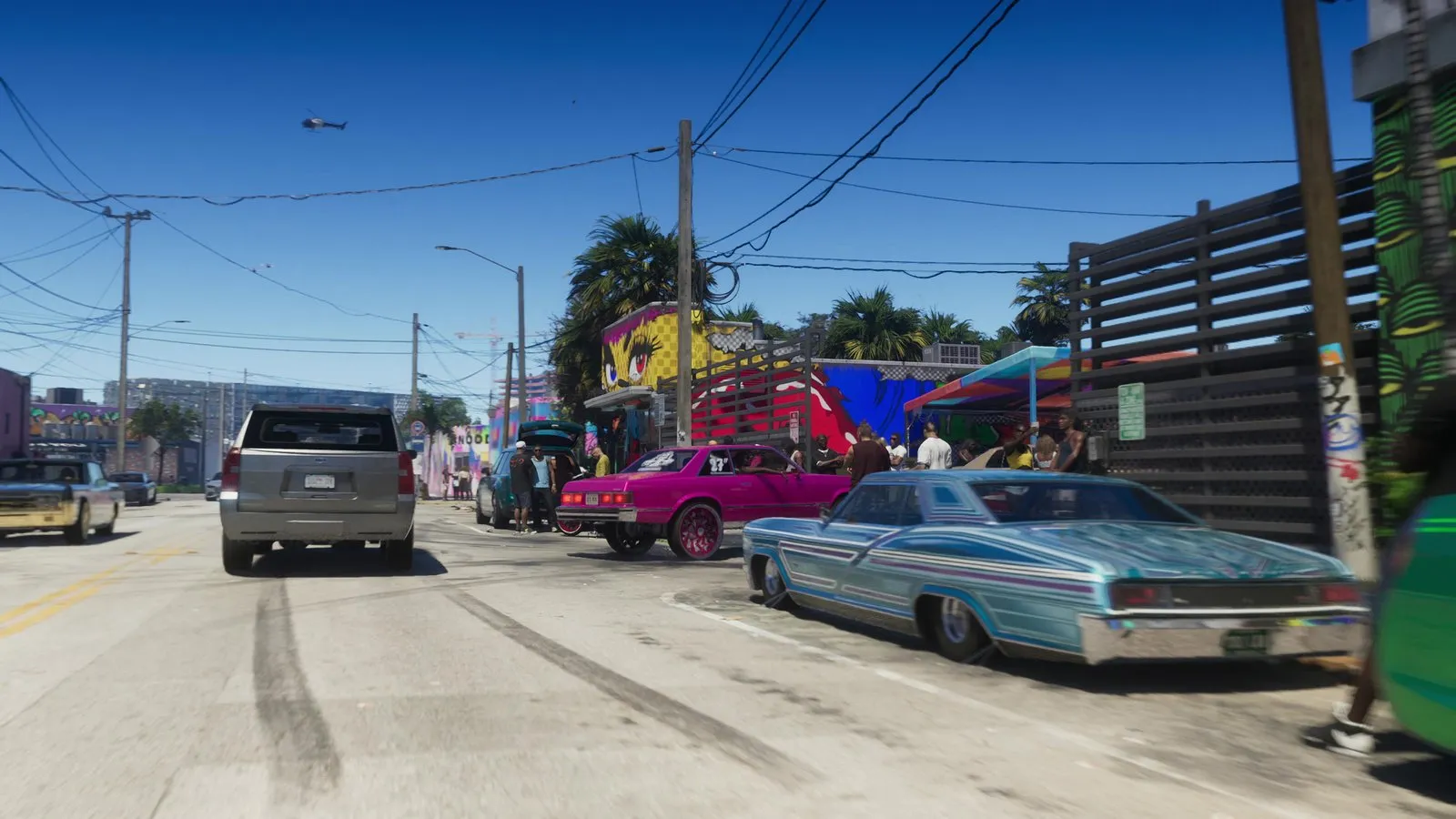 A distant helicopter flies over a mural lined street where a crowd gathers around lowriders and fresh tire marks.