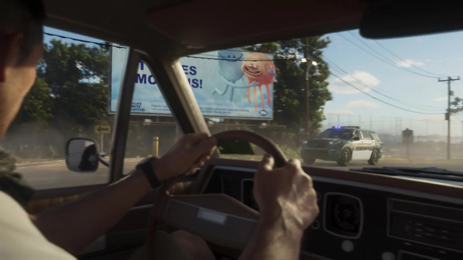 A sheriff SUV with blue lights on pulls onto the road ahead of a man driving an older car past a billboard.