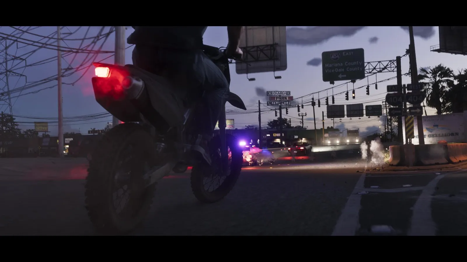 A dirt bike rider heads toward an intersection at dusk where a police vehicle with flashing lights sits amid gunfire and debris.